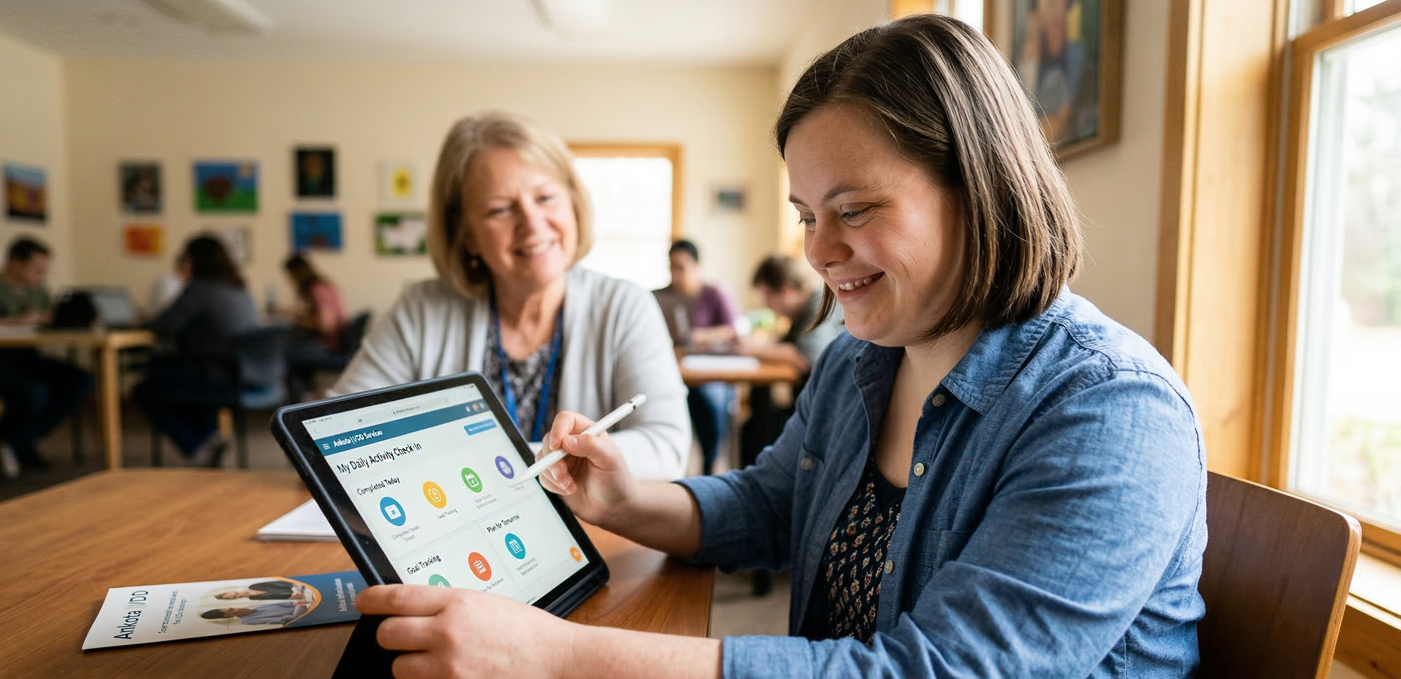 A young woman with Down syndrome sits at a wooden table, smiling as she interacts with a tablet using a stylus. The screen shows a "Daily Activity" dashboard with colorful icons. In the background, an older woman sits supportively, smiling. An "Ankota I/DD" brochure is visible on the table.